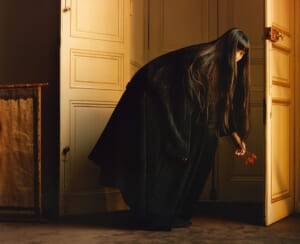 Woman with long black hair and a dark cloak bends down at a warmly lit doorway, holding a red flower near the floor.