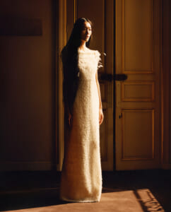 Woman in a long textured cream dress stands in a doorway, bathed in warm amber light.