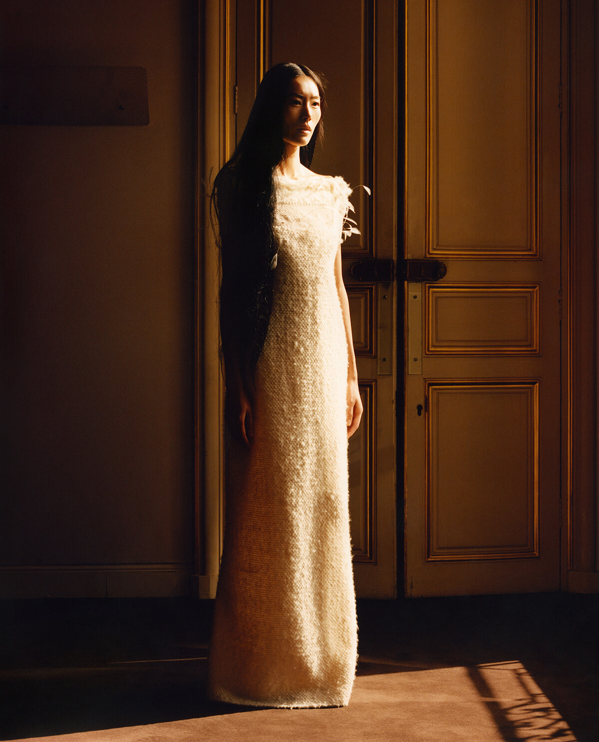 Woman in a long textured cream dress stands in a doorway, bathed in warm amber light.
