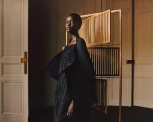 Model standing in a doorway wearing a dark pleated cape over a dress, with a wooden room divider behind.