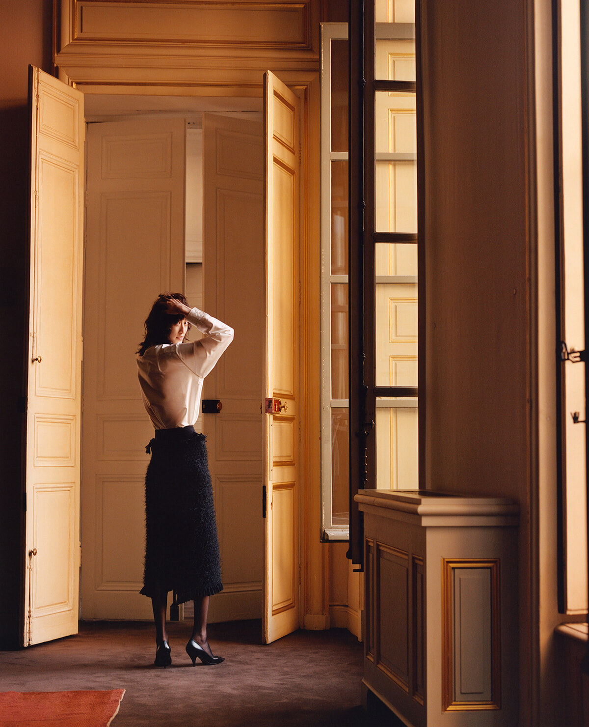 Woman in a white blouse and dark textured skirt adjusts her hair while standing in a doorway in a warmly lit, grand interior with tall doors and windows.