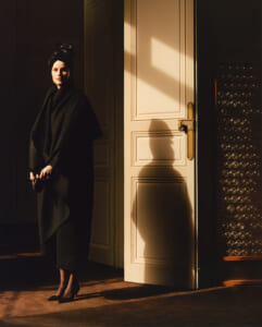 Woman in a black coat and headwrap stands by an open door, warm sunlight casting long shadows on the wall and floor