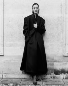 Fashion model stands against a light stone wall in an oversized black coat, hands at the lapel, wearing statement earrings.