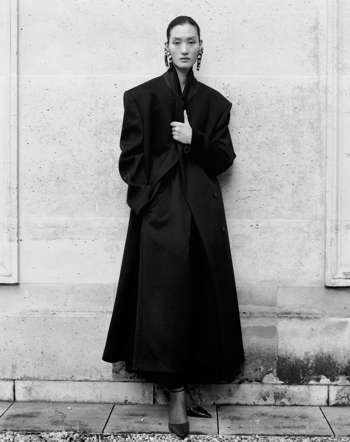 Fashion model stands against a light stone wall in an oversized black coat, hands at the lapel, wearing statement earrings.