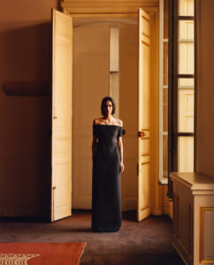Woman in an off-the-shoulder black gown standing in an open doorway of a warmly lit, classic interior.