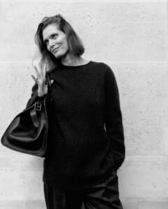 Black-and-white portrait of a woman in a dark sweater, holding a large black bag and waving at the camera against a concrete wall.