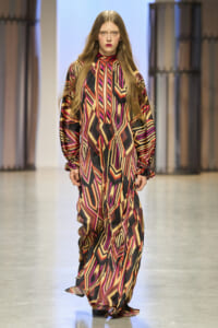 Model walking the runway in a long geometric-patterned dress with bold red, pink, orange, and black zigzags.