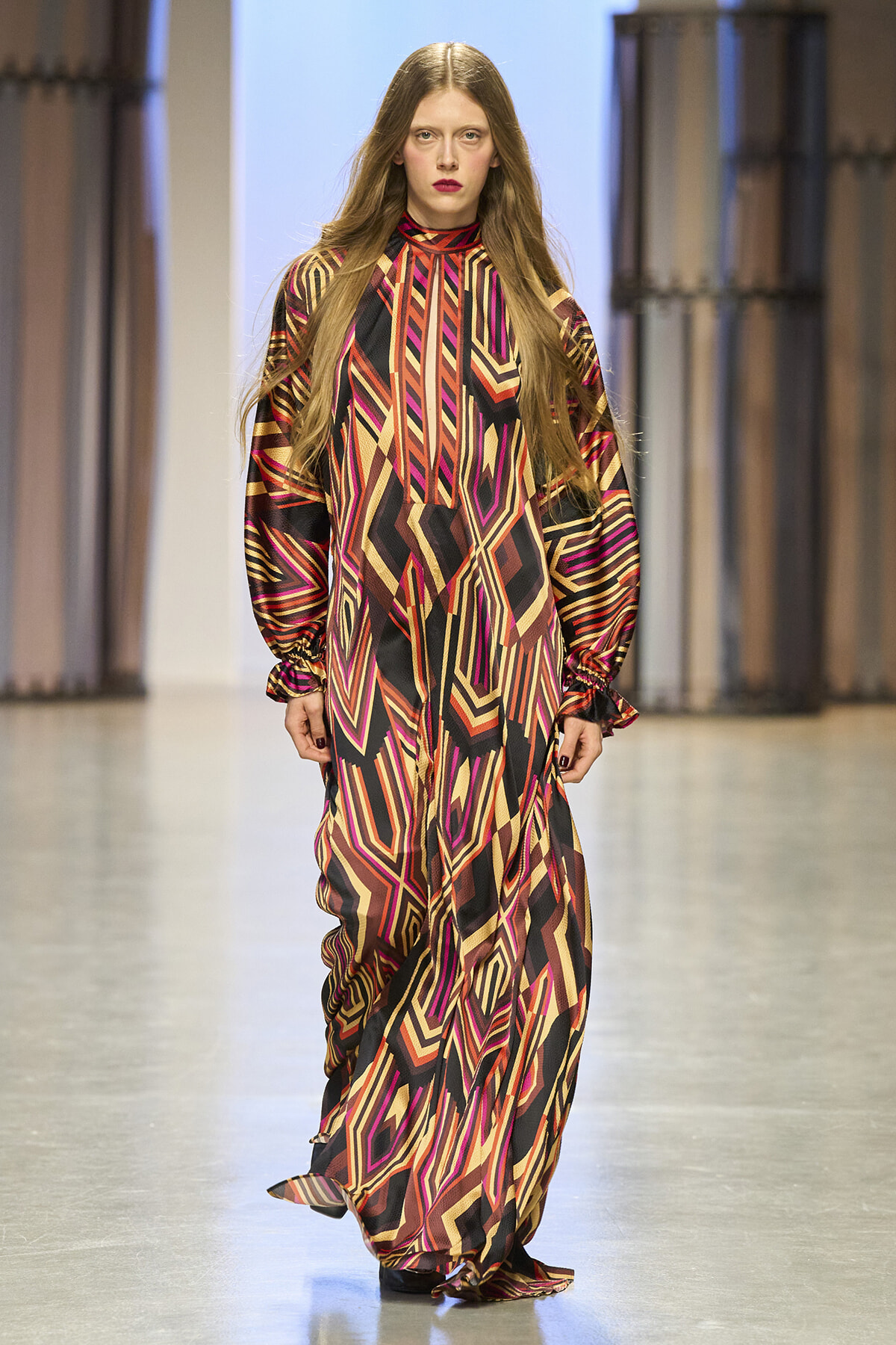 Model walking the runway in a long geometric-patterned dress with bold red, pink, orange, and black zigzags.
