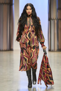 Model walks the runway in a bold geometric-print jumpsuit in red, orange, gold, and black, with long black hair and knee-high boots, carrying a matching patterned scarf bag.