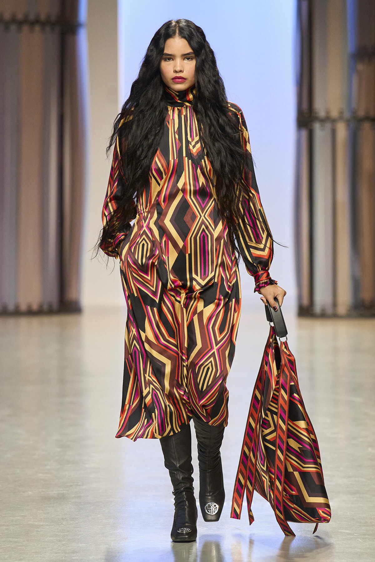 Model walks the runway in a bold geometric-print jumpsuit in red, orange, gold, and black, with long black hair and knee-high boots, carrying a matching patterned scarf bag.