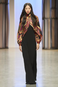 Model walking the runway in a bold multicolored geometric cape over black wide-leg pants.