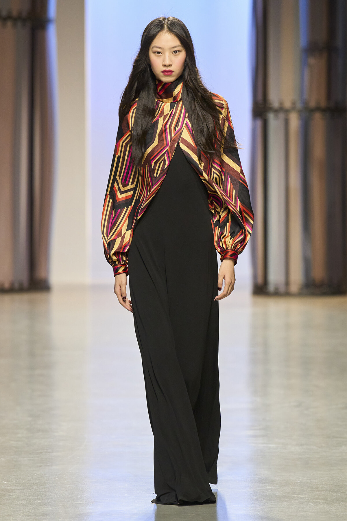 Model walking the runway in a bold multicolored geometric cape over black wide-leg pants.