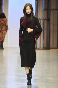 Model walking on a runway in a black turtleneck and midi black skirt, holding a burgundy and black geometric wrap across her torso.