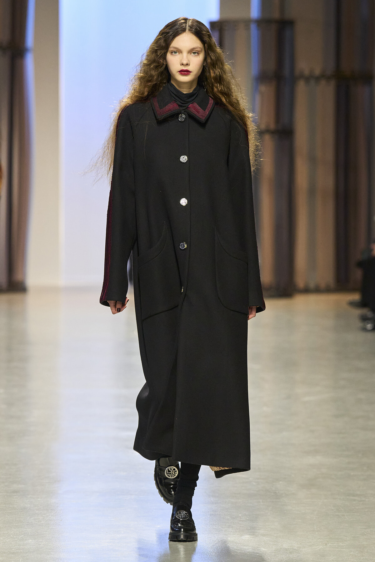 Female model walking a fashion runway in a long black coat with burgundy collar and silver buttons.