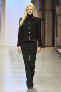 Female model walking a runway in a black tailored jacket with red trim and silver buttons, paired with black trousers.