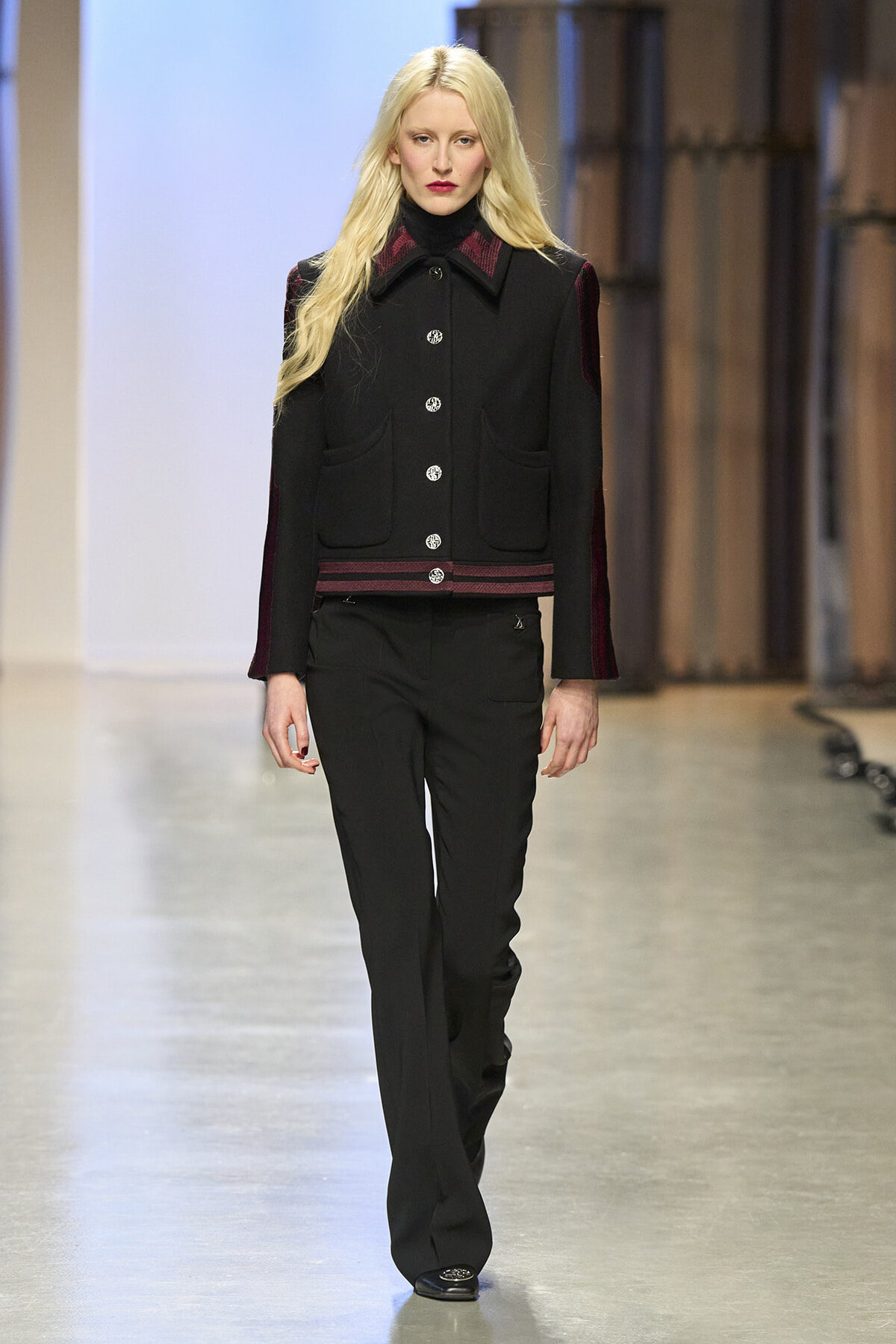 Female model walking a runway in a black tailored jacket with red trim and silver buttons, paired with black trousers.
