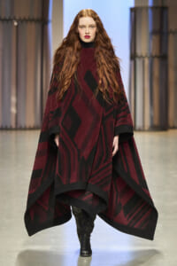 Model walking a runway in a burgundy and black geometric poncho with black trim, paired with a black turtleneck and tall boots.
