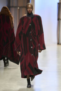 Model walking the runway in a long burgundy and black geometric-patterned coat with large buttons.