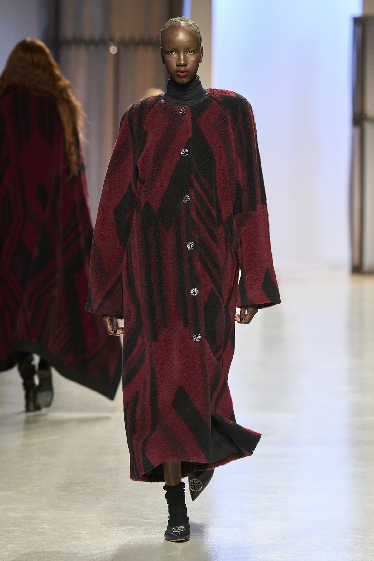 Model walking the runway in a long burgundy and black geometric-patterned coat with large buttons.