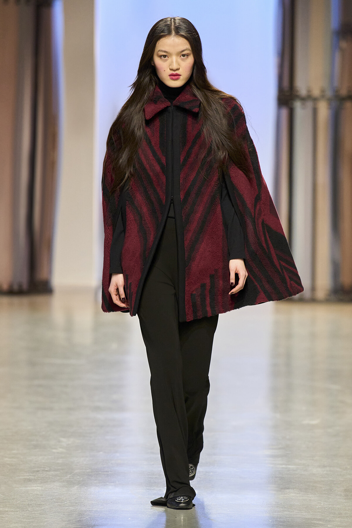 Model walking the runway in a burgundy and black patterned cape over a black outfit.