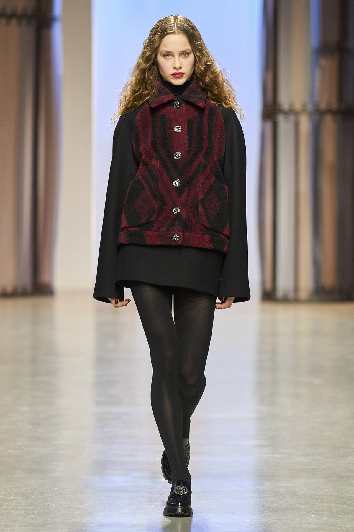 Model walks the runway in a red and black plaid vest over a black layered dress, black tights, and loafers.