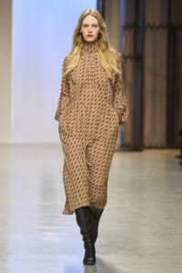 Model walking a runway in a tan patterned jumpsuit with a high neck, long sleeves, and black knee-high boots.