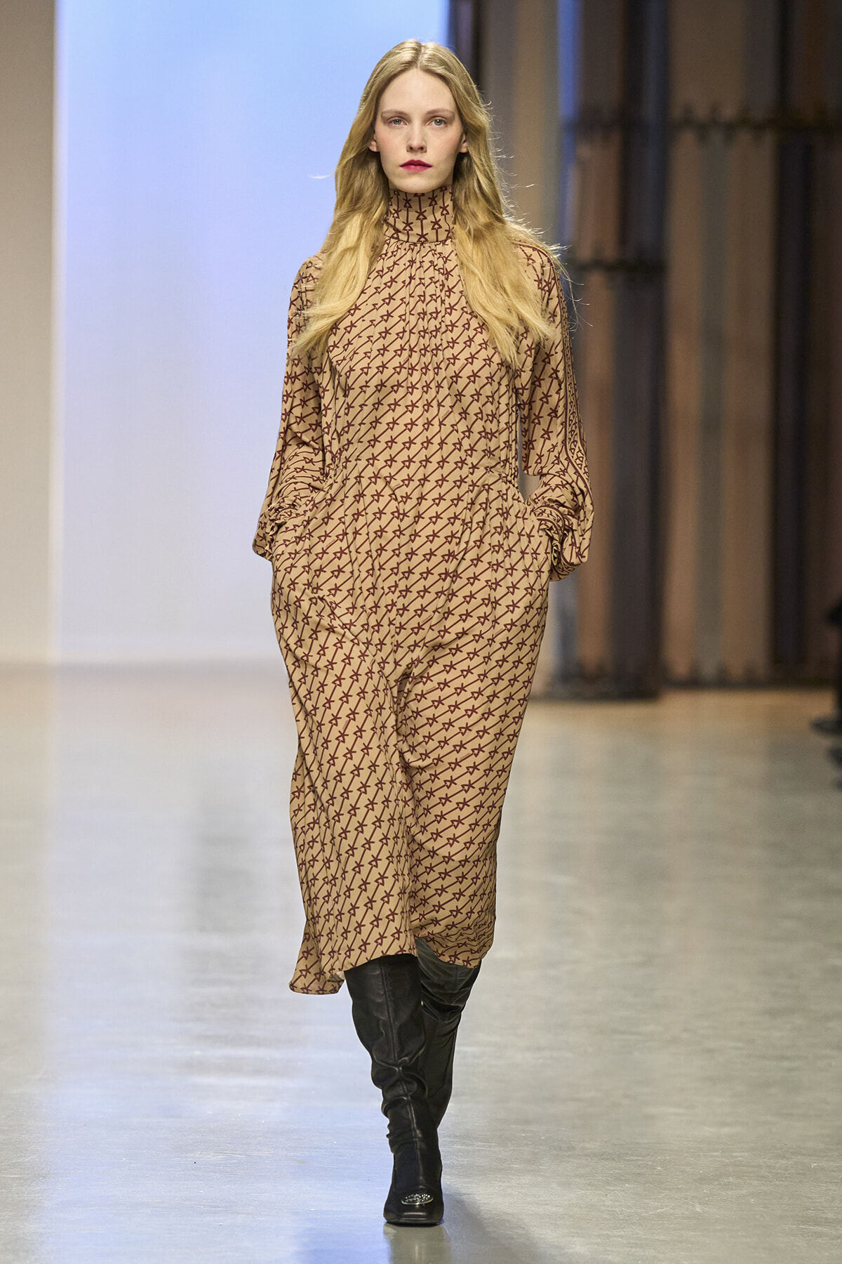 Model walking a runway in a tan patterned jumpsuit with a high neck, long sleeves, and black knee-high boots.