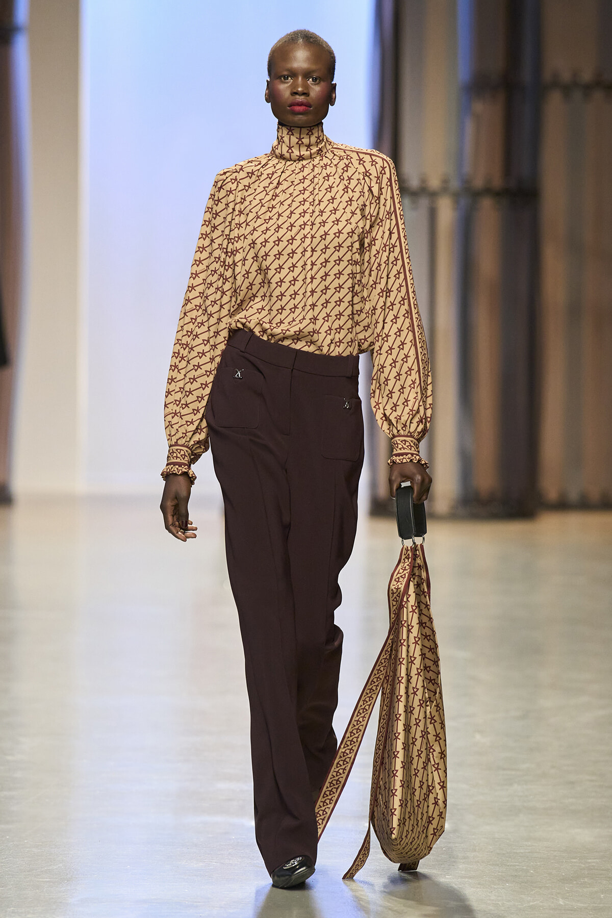 Model walks a fashion runway in a beige patterned turtleneck blouse and dark brown trousers, holding a matching scarfed bag at her side.