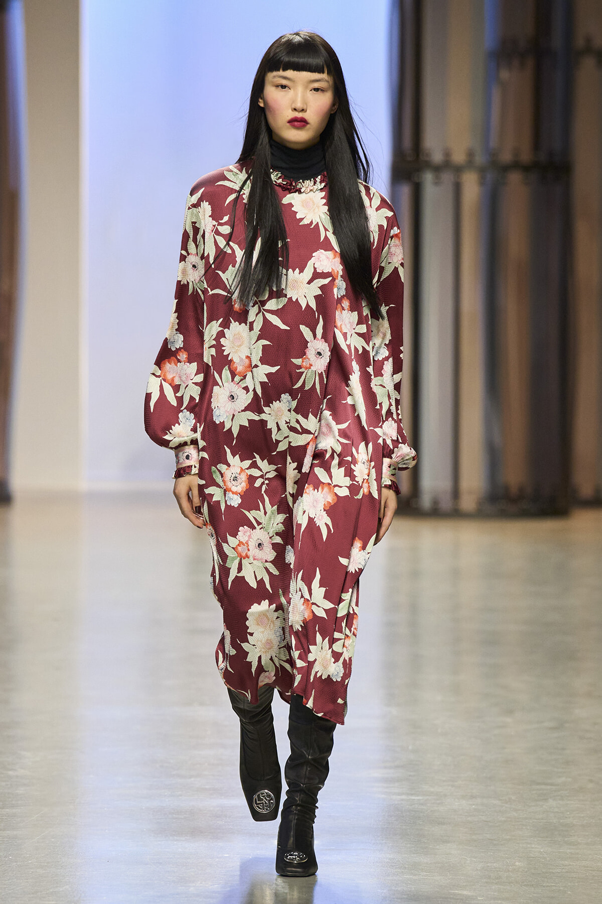 Model walking a runway in a burgundy floral dress over a black turtleneck, with knee-high black boots.