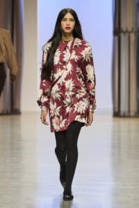 Model walking a runway in a burgundy floral dress with black tights and flats, on a fashion show catwalk.