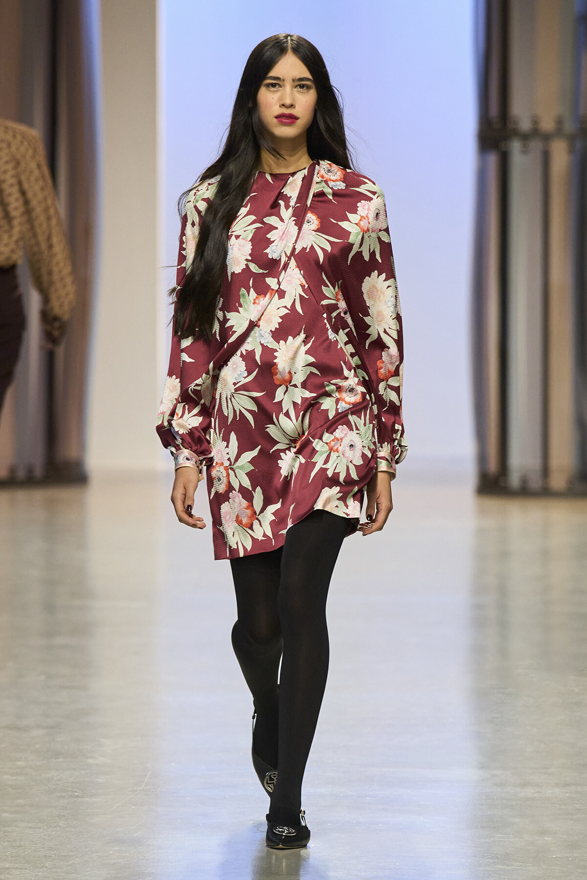 Model walking a runway in a burgundy floral dress with black tights and flats, on a fashion show catwalk.
