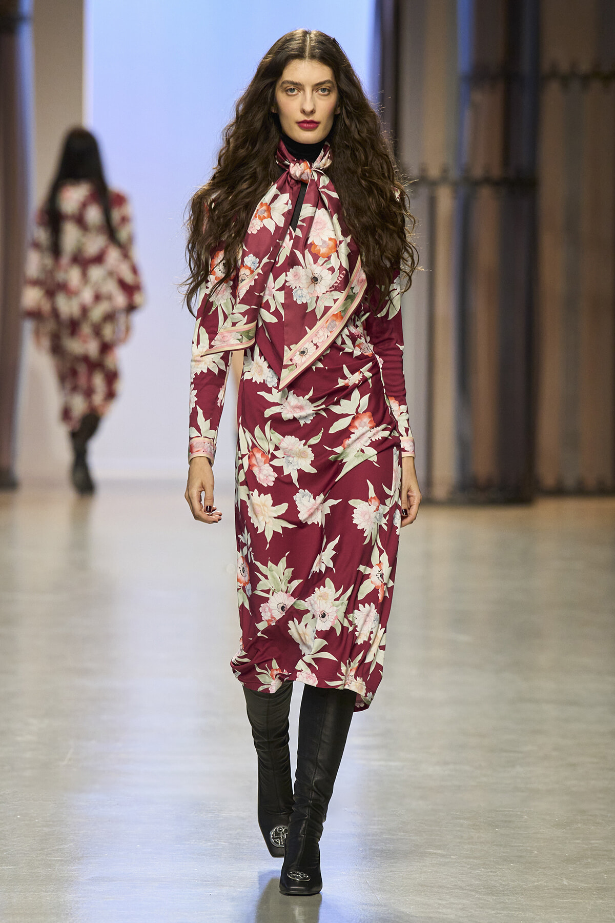 Model walking a fashion runway in a burgundy floral dress with a large scarf bow and black knee-high boots.