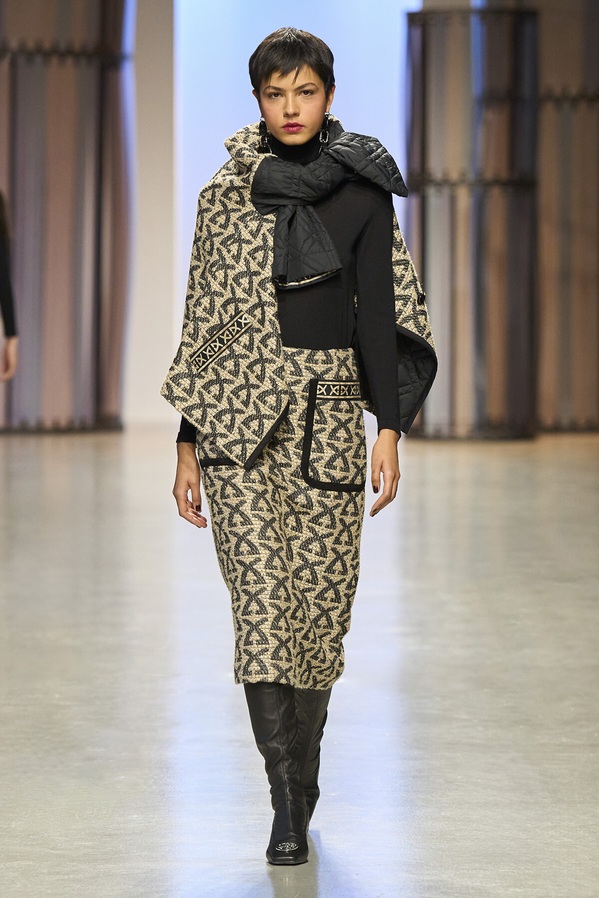 Model on a runway wearing a beige and black geometric patterned suit with a black top and knee-high boots, cape-like outerwear draped over shoulders.