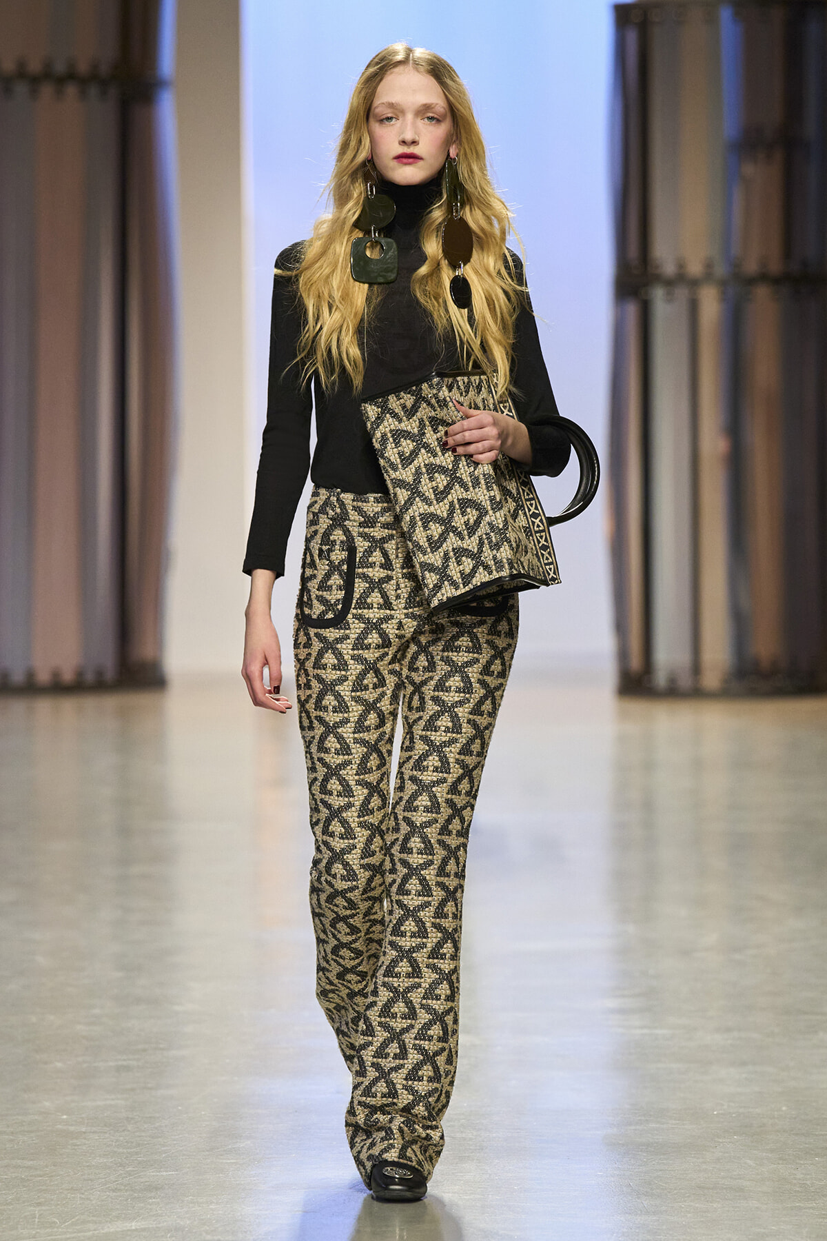 Model walking runway in a black turtleneck and patterned pants, carrying a matching geometric clutch with circular handle and oversized earrings.