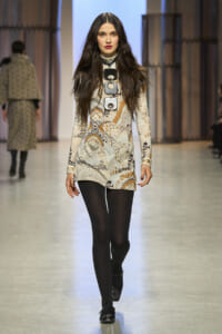 Model walking a fashion show runway in a beige-brown patterned long-sleeve mini dress, black tights, and black loafers, with a large rectangular necklace centerpiece.