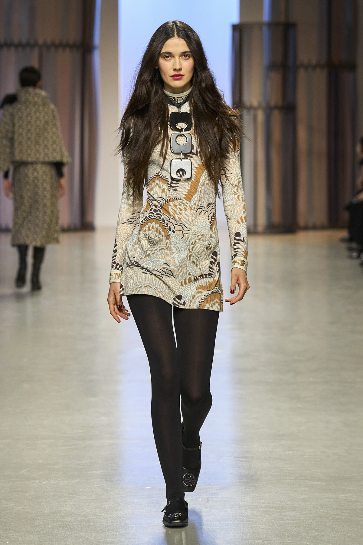 Model walking a fashion show runway in a beige-brown patterned long-sleeve mini dress, black tights, and black loafers, with a large rectangular necklace centerpiece.