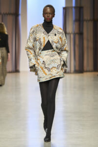 Model walking a fashion runway wearing a loose, patterned coat over a black turtleneck and tights; bold orange, blue, and beige print with a black top underneath.