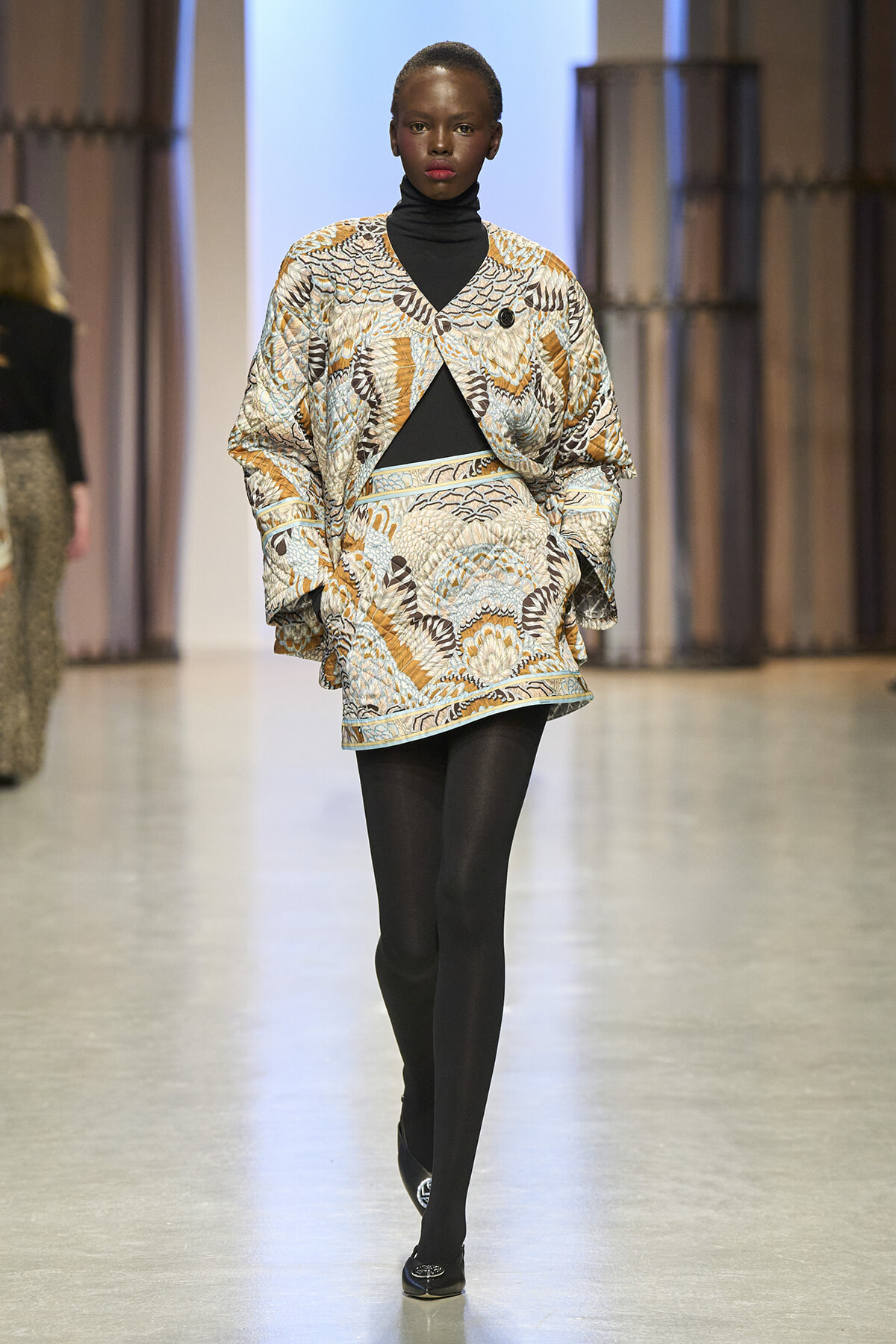 Model walking a fashion runway wearing a loose, patterned coat over a black turtleneck and tights; bold orange, blue, and beige print with a black top underneath.