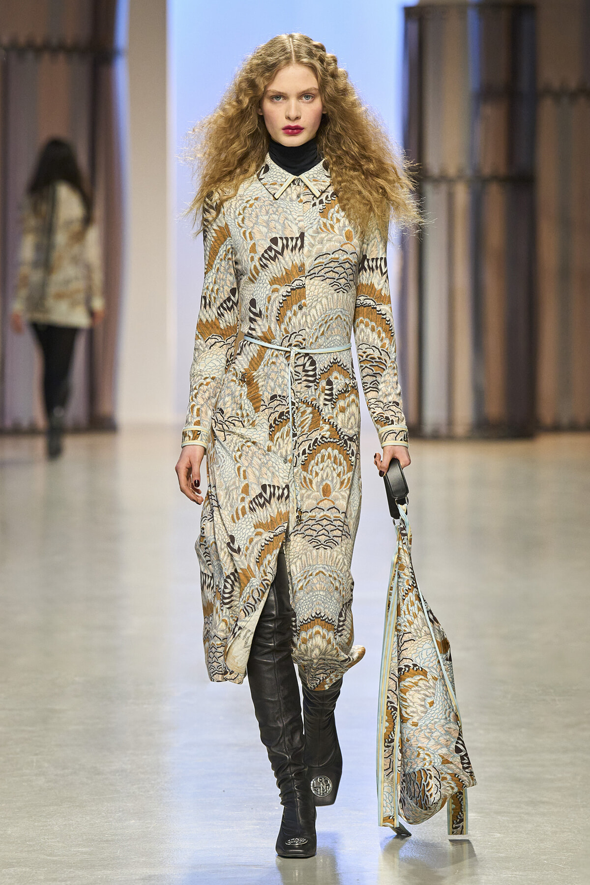 Female model walks a fashion runway wearing a long patterned coat with a belt, black turtleneck and knee-high boots.