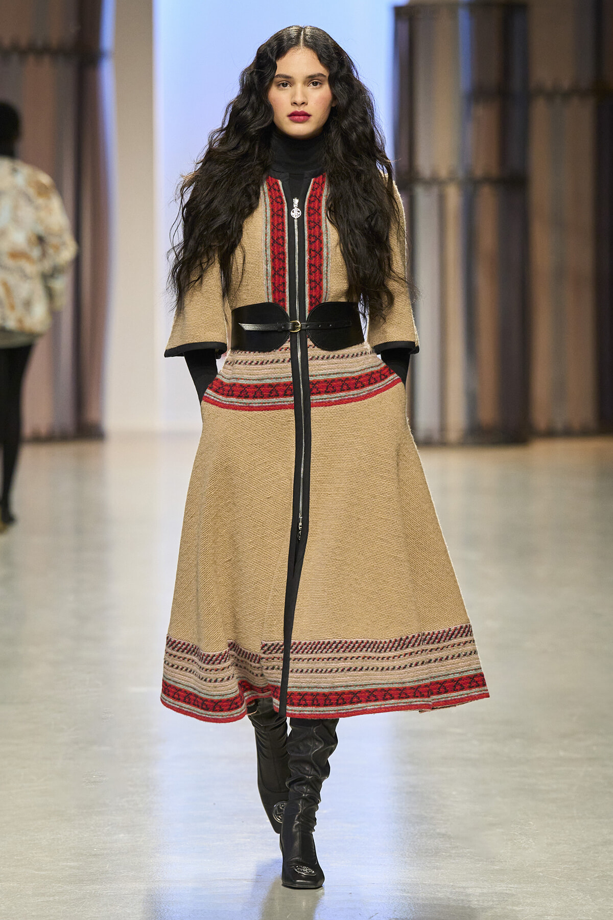 Fashion model walking runway in beige knit coat with red and black patterned trim, black turtleneck, belt, and black knee-high boots.