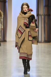 Model walks the runway in a camel-colored knit poncho with red patterned trim and matching skirt, paired with black boots and a beige bag with black handles.
