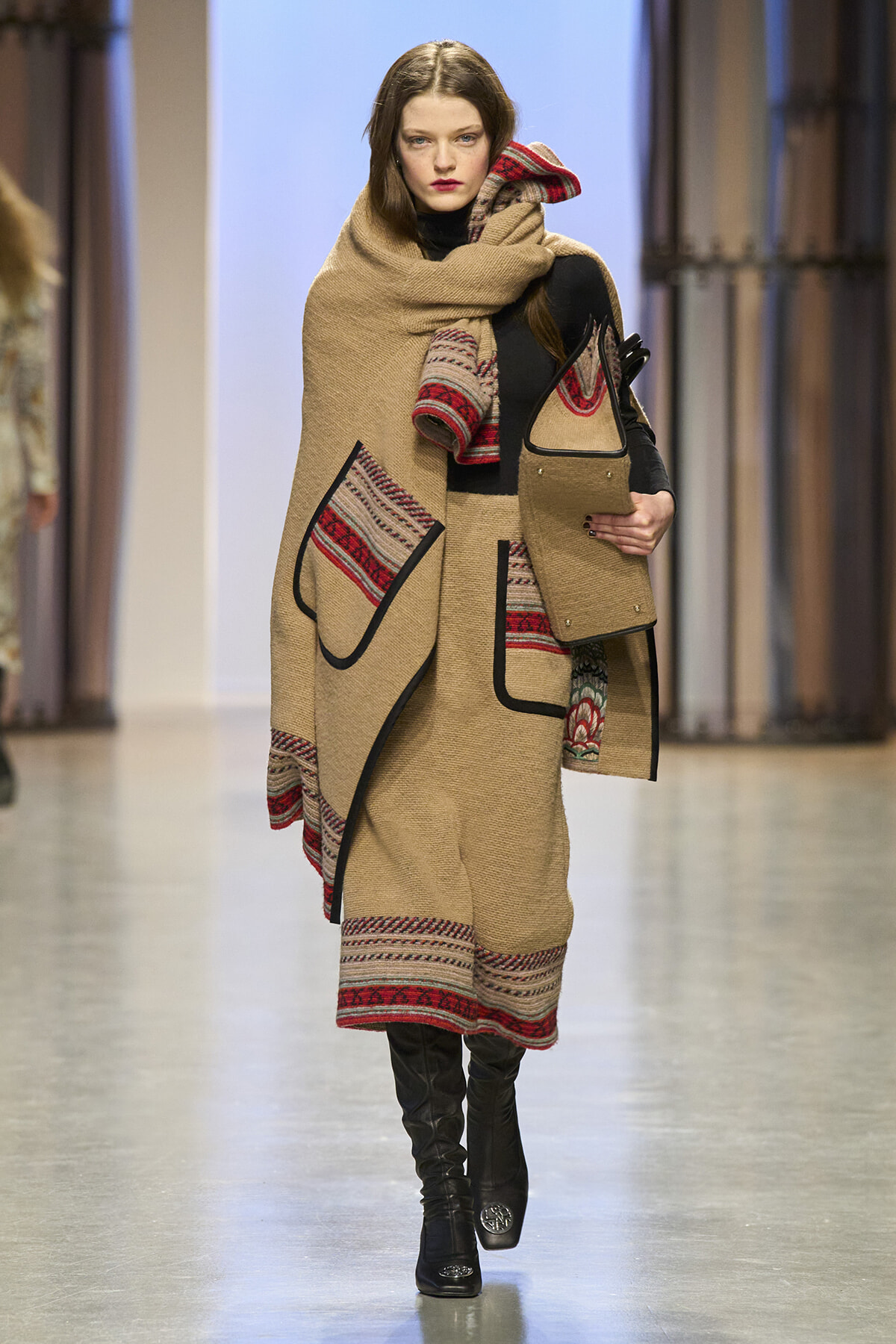 Model walks the runway in a camel-colored knit poncho with red patterned trim and matching skirt, paired with black boots and a beige bag with black handles.