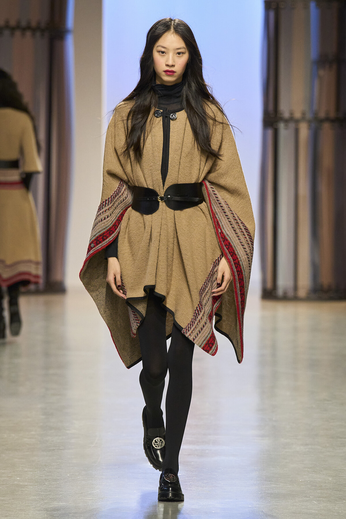 Model walking a runway in a camel poncho with red patterned trim, black belt, and black tights with loafers on a fashion show catwalk.
