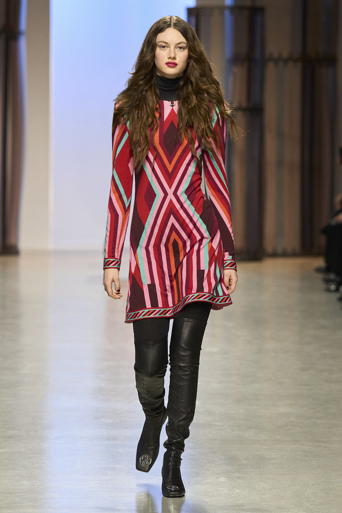 Model walking a runway in a bold geometric-patterned dress in pink, red, and teal with a black turtleneck under it and black thigh-high boots.