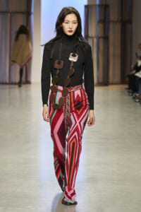 Model on runway wearing a black turtleneck with vivid pink-red geometric pants and a long necklace of large brown circular discs.