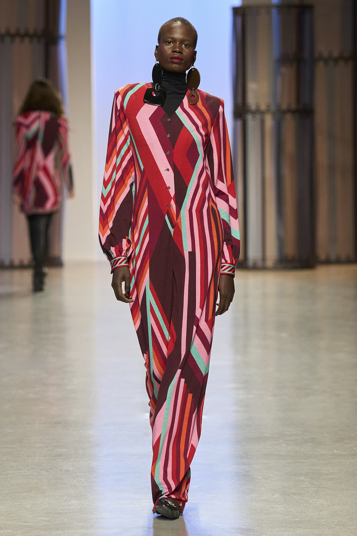 Model walking a fashion runway in a bold geometric print jumpsuit with red, pink, and teal stripes and oversized earrings.