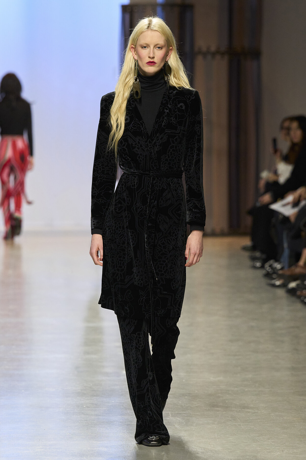 Model walking a runway in a black velvet patterned suit with a belt and turtleneck, long blonde hair and bold earrings.