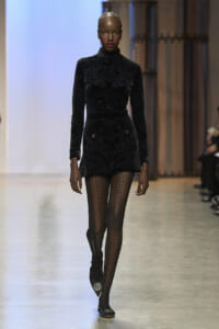 Model walking a fashion runway in a black velvet-patterned turtleneck mini dress with long sleeves, paired with fishnet tights and pointed heels.