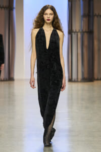Model walks down a runway in a black halter jumpsuit with a plunging V neckline and subtle floral pattern.