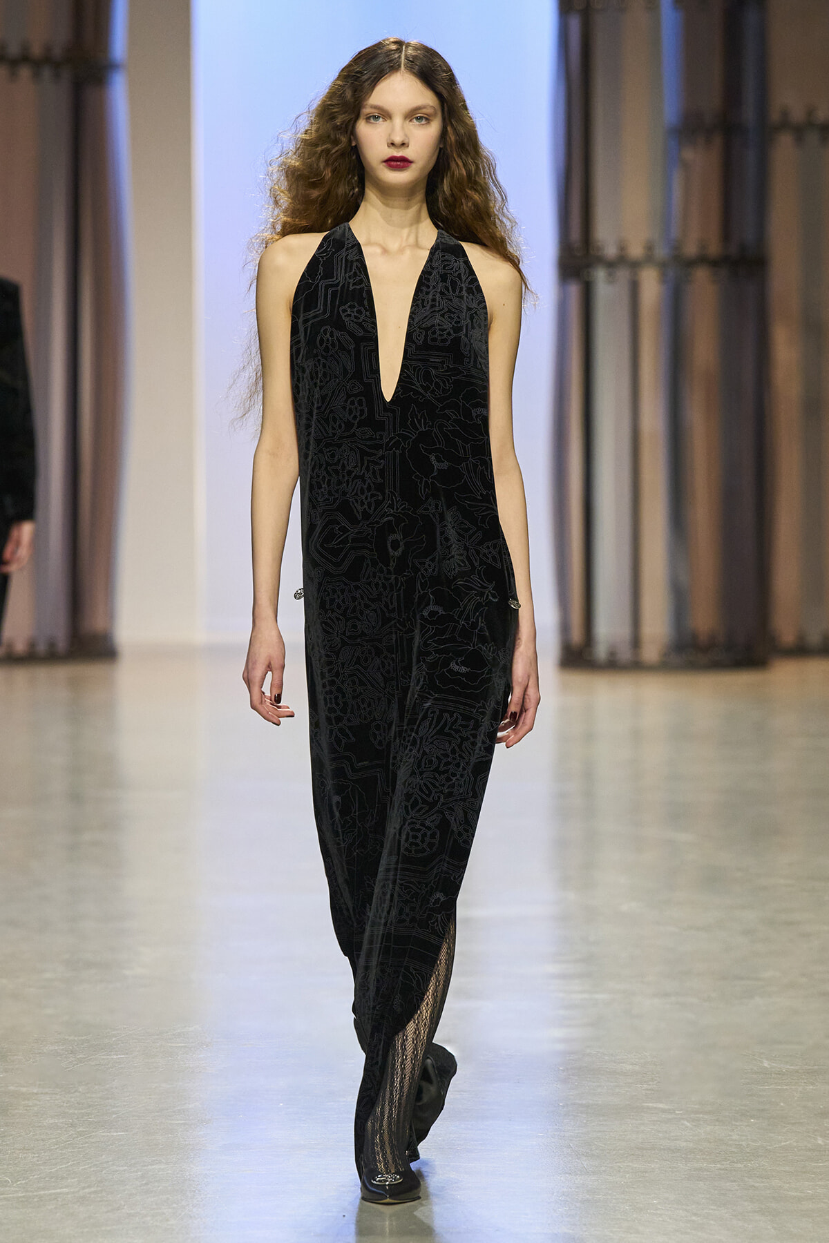 Model walks down a runway in a black halter jumpsuit with a plunging V neckline and subtle floral pattern.